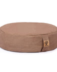 A round meditation cushion in a warm taupe-brown fabric, featuring a small sewn-on cork patch on the front right side, set against a plain white background