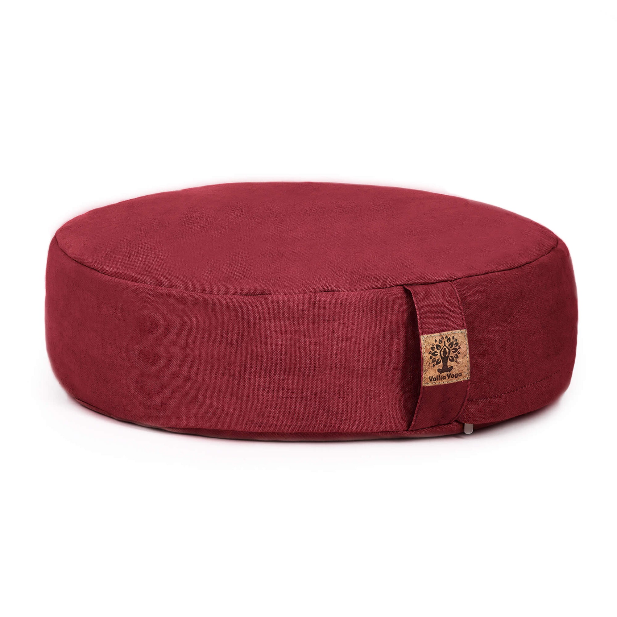A round, deep red fabric meditation cushion with a small cork tag on the side, photographed on a clean white background